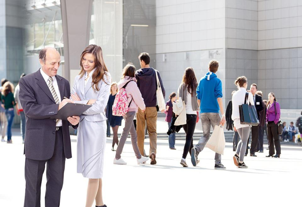 Two people in business attire review documents outside a modern glass building, possibly consulting an Asset Protection Attorney, while several casually dressed individuals walk by in the background.