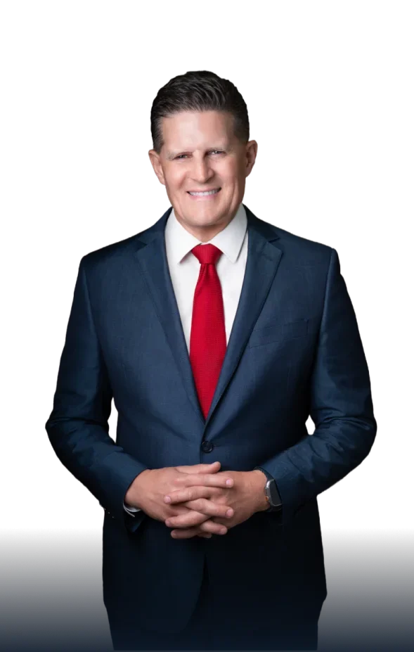 Man in a dark blue suit, white shirt, and red tie standing with hands clasped, smiling at the camera against a black background.