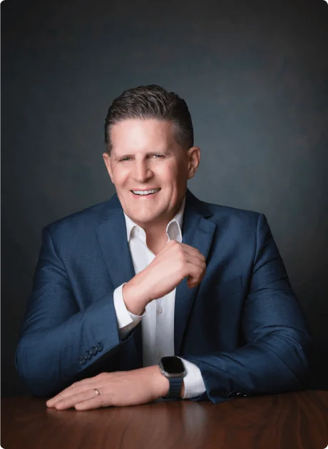 A man in a blue suit jacket and white shirt sits at a table, smiling at the camera with one hand resting on the table and the other adjusting his jacket.