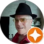 A man wearing a black hat, yellow-tinted glasses, and a red shirt is pictured indoors. An orange star badge icon in the lower right corner suggests he may be featured in highlighted testimonials.