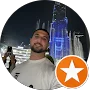 A man in a white shirt poses for a photo at night in front of a tall, illuminated building with blue lights and fountains; an orange star rating icon in the corner hints at glowing testimonials.
